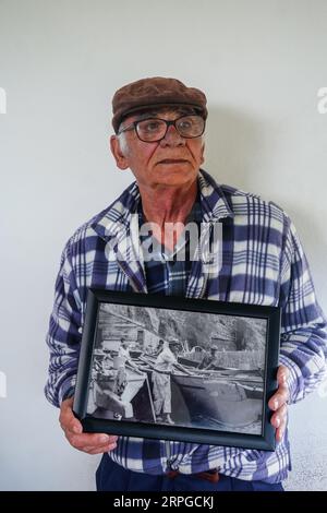 Luis Mariano, der letzte lebende Walfänger auf der Insel Sao Miguel, posiert mit einem Foto von seinen Walfangtagen an einer ehemaligen Walfangstation in der Nähe von Agua de Pau, Sao Miguel Island, Azoren, Portugal. Das Töten von Walen wurde 1986 auf den portugiesischen Inseln verboten und durch Walbeobachtungstouren ersetzt, die profitabler und nachhaltiger sind. Stockfoto