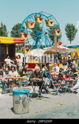 Die Leute genießen Essen an Tischen in der Nähe von Imbissständen beim Rollende Keukens Food Festival, mit einem blauen Riesenrad im Hintergrund Stockfoto