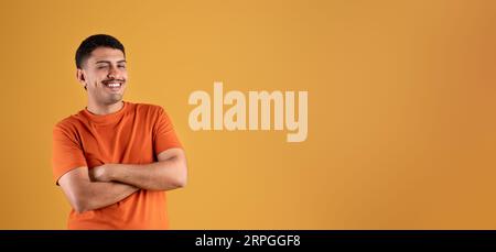 Positiver brasilianischer Mann in orangefarbenem T-Shirt mit gekreuzten Armen auf der Brust, blinkend und lächelnd, Panorama, freier Platz Stockfoto
