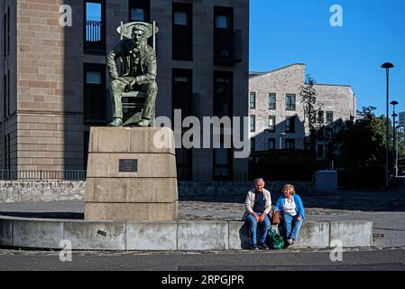 Ein älteres Paar nimmt neben der sitzenden Statue des schottischen Architekten und Designers Charles Rennie Mackintosh Platz, die von Andy Scott geformt wurde. Glasgow. Stockfoto