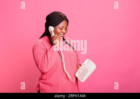 Porträt eines lächelnden afroamerikanischen Models, das auf dem Festnetztelefon chattet, während es im Studio mit rosafarbenem Hintergrund steht. Eine Frau, die sich mit einem stationären Vintage-Telefon aus der Ferne unterhält Stockfoto