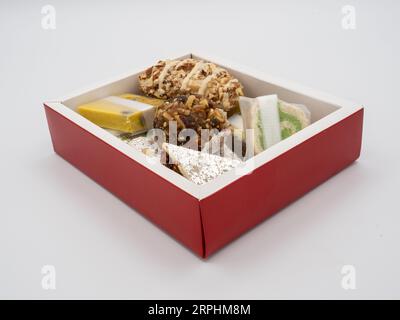 Winkelansicht einer Box mit roten Seiten, gefüllt mit verschiedenen, köstlichen indischen Ghee-Süßigkeiten, isoliert auf weißem Hintergrund. Stockfoto