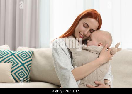 Mutter mit Spielzeughase, die ihr Kind zu Hause in einer Schlinge (Babytrage) hält Stockfoto