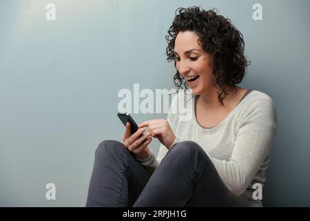 Frau auf der Etage ihres Zuhauses, die von den digitalen Inhalten ihres Smartphones begeistert ist. Von Videos bis hin zu sozialen Medien ist jede Unterhaltungsform zugänglich. Donni Stockfoto