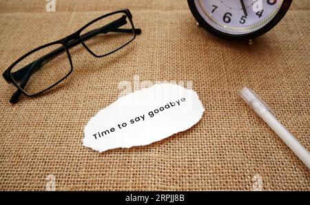 Eine Wecker-Uhr, Gläser, Stift und ein zerrissenes Stück Cremepapier, das Zeit zum Abschied sagt! Auf einem Sackleinen-Hintergrund. Stockfoto