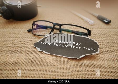 Eine Wecker, Gläser, Stift und ein zerrissenes Stück Cremepapier, das mit Menschen geschrieben wurde, kaufen von Menschen, denen sie vertrauen, auf hölzernem Hintergrund Stockfoto