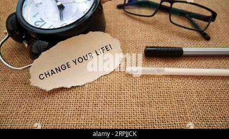 Eine Wecker-Uhr, eine Brille, ein Stift und ein zerrissenes Stück Cremepapier, auf dem steht „Change Your Life!“. Auf einem Sackleinen-Hintergrund. Stockfoto
