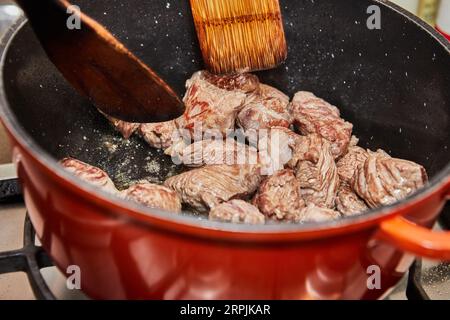 Rindfleischstücke werden in einer Pfanne auf einem Gasherd gebraten. Holzstäbchen zum Rühren Stockfoto