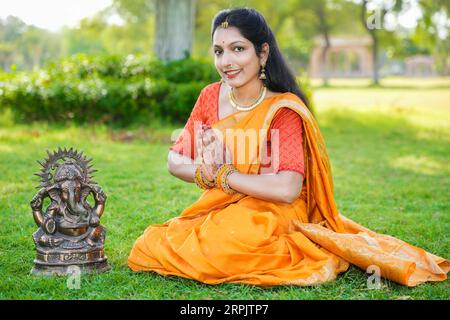 Glückliche junge indische Frau, die Saree trägt, die im Park, der das Ganesh Chaturthi Festival feiert, sitzt und Lord Ganesha Idol anbetet. Stockfoto