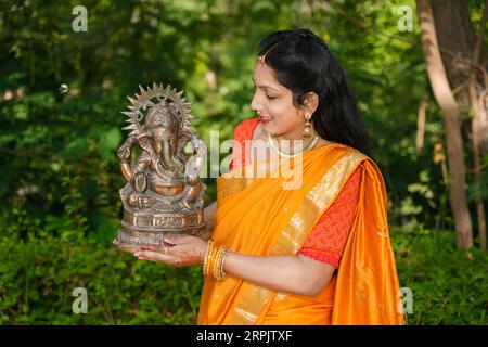 Glückliche junge indische Frau trägt Saree und hält Lord Ganesha Idol im Freien im Park, der Ganesh Chaturthi Festival feiert. Stockfoto