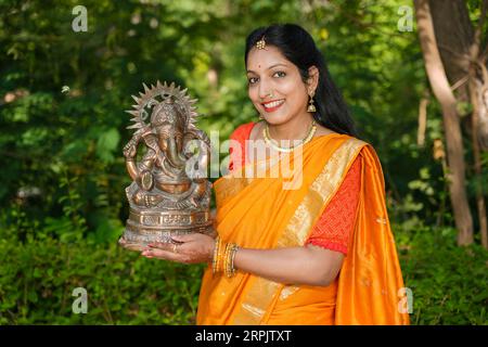 Glückliche junge indische Frau trägt Saree und hält Lord Ganesha Idol im Freien im Park, der Ganesh Chaturthi Festival feiert. Stockfoto