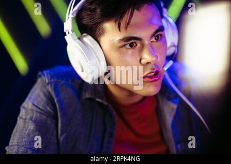 Junger männlicher Gamer, der mit fixierten Augen auf dem Computerbildschirm vor ihm sitzt. Wenn er ein Headset trägt, ist sein Ausdruck angespannt und seine Augenbrauen sind f Stockfoto