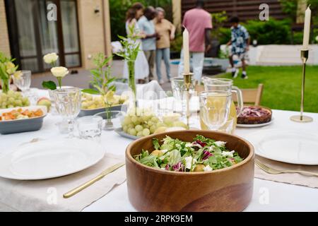 Konzentrieren Sie sich auf eine Holzschale mit frischem, hausgemachtem vegetarischen Salat, der auf dem Serviertisch zwischen Tellern, Gabeln, Obst, Kerzen, Gläsern und Blumen steht Stockfoto