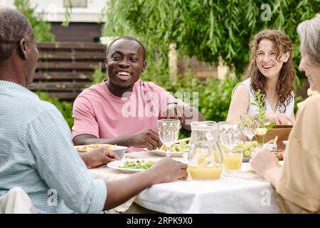 Glückliches junges interkulturelles Paar, das beim Gespräch mit den Eltern beim Abendessen lacht, während es am Tisch sitzt und mit hausgemachten Speisen und Getränken serviert wird Stockfoto