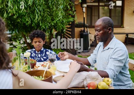 Reifer afroamerikanischer Mann und sein Enkel mit geschlossenen Augen, die Hände haltend und betend, während sie am servierten Tisch beim Abendessen sitzen Stockfoto