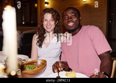 Glücklicher junger interkultureller Mann und Frau in Casualwear sitzt am servierten Tisch und schaut auf die Kamera, während er ein Familienessen im Freien genießt Stockfoto