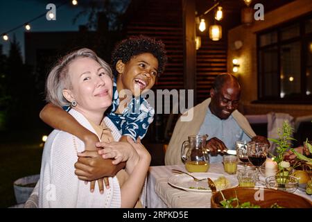 Glückliche reife Frau schaut in die Kamera, während ihr süßer Enkel sie beim Familienessen im Hinterhof am Tisch begrüßt Stockfoto