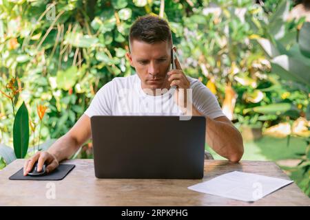 Software-Designer, der ein neues Projekt mit seinem Kunden am Telefon diskutiert. Seriöser Mann, der in einem offenen Büro an einem Laptop arbeitet. Stockfoto