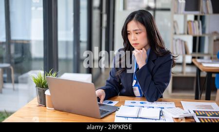 Eine gestresste und verärgerte junge asiatische Geschäftsfrau oder Finanzassistentin schaut mit einem besorgten und nachdenklichen Gesicht auf ihren Laptop-Bildschirm, Conce Stockfoto