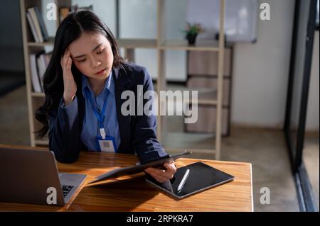 Eine nachdenkliche und fokussierte asiatische Geschäftsfrau oder Managerin in einem formellen Business-Anzug fühlt sich müde von Überlastung, während sie Dokumente liest Stockfoto