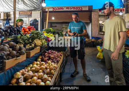 Zwei Männer stehen zwischen Kisten mit Gemüse auf dem überdachten Bauernmarkt in der Chavchavadze-Allee in Telavi, Georgien. Giorgi Chachua in einem türkisfarbenen T-Shirt mit hochgeschobenen Sonnenbrillen betrachtet ein kleines schwarzes Objekt in seiner Hand. Neben ihm steht ein junger Mann mit einer Kappe, Brille und Bart, der eine gelbe Plastiktasche trägt. Zwiebeln, braune Knollen, Auberginen, Tomaten, Blumenkohl, Brokkoli und Paprika liegen im Vordergrund. Links steht eine Marktverkäuferin mit einem grünen Kopftuch hinter ihrem Stand. Das Schild über einer Verkaufsnische im Hintergrund trägt die georgische Inschrift „ყავის დაფქვა“. Stockfoto