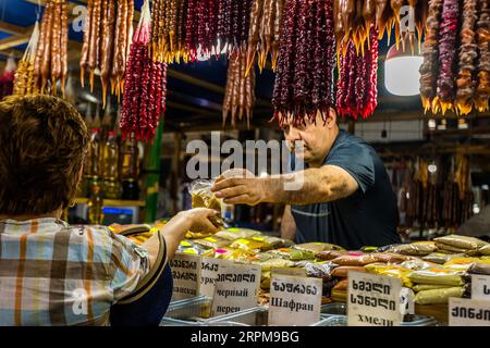 Ein Marktverkäufer überreicht einem Kunden in einem karierten Hemd eine Tüte Gewürz über die Theke seines Gewürzstandes auf dem überdachten Bauernmarkt in der Chavchavadze-Allee in Telavi, Georgien. Vor ihm stehen Plastiktabletts, dahinter befinden sich Gewürze in Tüten. Schilder auf Georgisch und Russisch nennen Svanetischer Salz (Georgisch სვანური მარილი), möglicherweise Kurkuma, schwarzen Pfeffer (შავი პილპილი), Safran (ზაფრანა) und Khmeli Suneli (ხმელი სუნელი). Svanetisches Salz kommt aus der Bergregion Svaneti. Neben Salz enthält es gewöhnlich Knoblauch, Koriandersamen, blauen Koriander, getrocknete Ringelblumen und getrocknete Chiliflügel. Khmeli Stockfoto