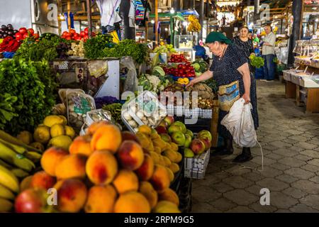 Eine ältere Frau mit einem grünen Kopftuch und einem gepunkteten Kleid greift mit ihrer rechten Hand in eine Kiste mit Kartoffeln an einem Gemüsestand auf dem überdachten Bauernmarkt in der Chavchavadze Avenue in Telavi, Georgien. Zwiebeln liegen daneben. In ihrer linken Hand hält sie eine weiße Plastiktüte, und eine Stofftasche hängt von ihrer Schulter. Pfirsiche, Äpfel, gelbe Früchte, Bananen, Knoblauch und Champignons in Schalen liegen im Vordergrund. Kräuterbüschel stehen links. Im Hintergrund stehen eine Frau in einem dunklen Kleid und ein Mann in einem hellen Hemd zwischen den Ständen. Ein gewölbtes Dach bedeckt den Hauptteil des Mar Stockfoto