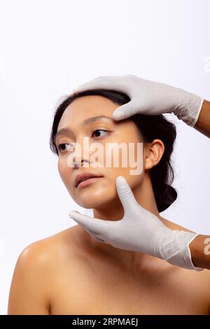 Hände der Kosmetikerin in weißen chirurgischen Handschuhen, die das Gesicht einer asiatischen Frau mit dunklem Haar halten, Kopierraum Stockfoto