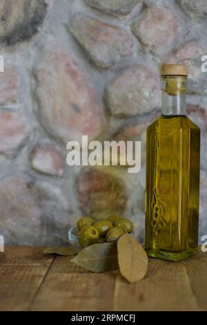 Eine Glasflasche Olivenöl steht auf einem Holztisch mit Oliven und einer strukturierten Steinmauer im Hintergrund, die ein rustikales Ambiente schafft. Stockfoto