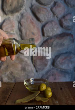 Eine Glasflasche Olivenöl steht auf einem Holztisch mit Oliven und einer strukturierten Steinmauer im Hintergrund, die ein rustikales Ambiente schafft. Stockfoto