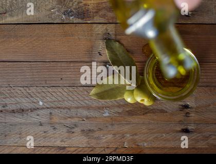 Eine Glasflasche Olivenöl steht auf einem Holztisch mit Oliven und einer strukturierten Steinmauer im Hintergrund, die ein rustikales Ambiente schafft. Stockfoto