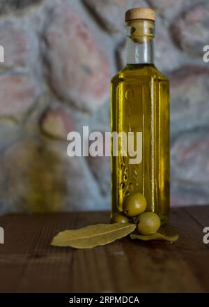 Eine Glasflasche Olivenöl steht auf einem Holztisch mit Oliven und einer strukturierten Steinmauer im Hintergrund, die ein rustikales Ambiente schafft. Stockfoto