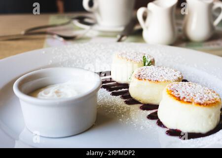Hüttenkäse-Pfannkuchen mit Kirschmarmelade und Sauerrahm. Vanillesyrniki auf weißem Teller. Süßer Quark Dessert. Süßes Frühstückskonzept. Stockfoto
