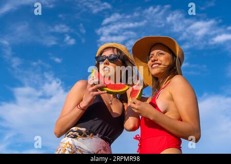 Porträt weiblicher Freunde am Strand im Sommer lächelnd beim Essen einer Wassermelone mit dem Himmel im Hintergrund, Bottom-up-Aufnahme Stockfoto