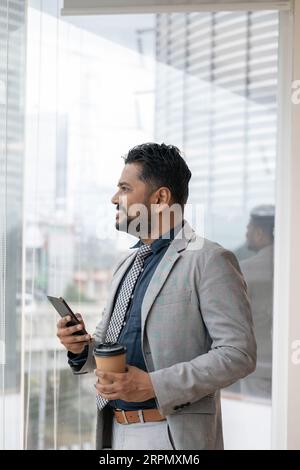 Erfolgreicher Geschäftsmann indianer arbeitet im Büro bei der Arbeit, Geschäftsmann indianer im Geschäftsanzug verwendet Telefon in der Nähe des Fensters, Mann lächelt und liest gut Stockfoto