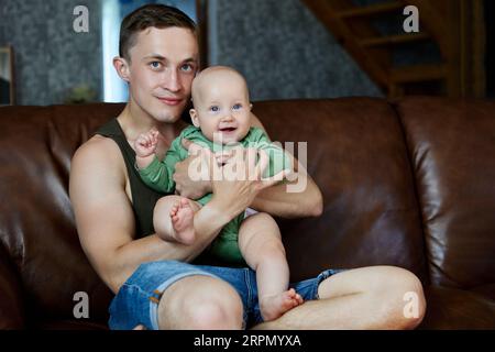 Junger Vater 26 Jahre alt mit Tochter im Arm sitzt auf Sofa im Wohnzimmer, Familienportrait. Stockfoto