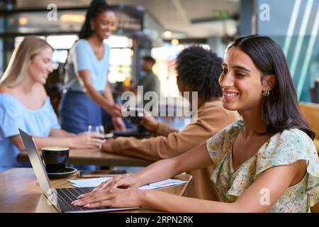 Frau, Die Am Laptop Arbeitet, Sitzt Am Tisch In Einem Geschäftigen Café Stockfoto