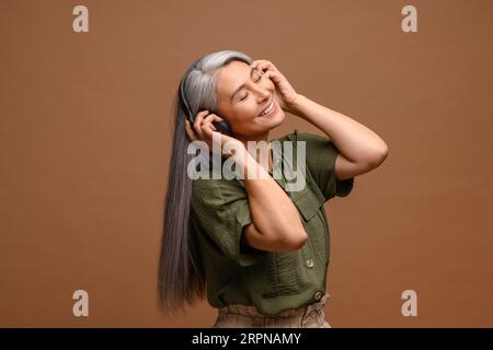 Eine ruhige asiatische Frau mittleren Alters in Kopfhörern, die Musik hört und isoliert auf braunem Hintergrund tanzt, eine entspannte, grauhaarige Dame aus den 50er Jahren, die Lieblingslieder genießt Stockfoto