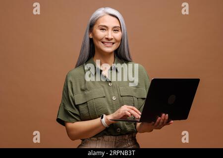 Grauhaarige asiatische Geschäftsfrau der 40er Jahre der 50er Jahre, die ein Notebook einsetzt, Schülerinnen mittleren Alters, die schreiben, fröhliche Frau, die auf E-Mails reagiert, Werbekonzept Stockfoto