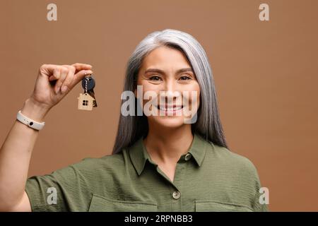 Asiatische Frau mittleren Alters, die Schlüssel isoliert auf braunem Hintergrund zeigt, fröhliche lächelnde grauhaarige Dame aus den 50er Jahren, die Immobilien kauft Stockfoto