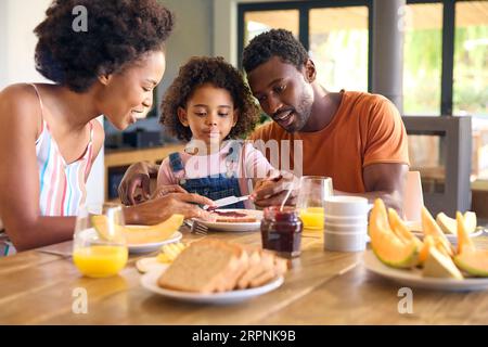 Familienschoß Mit Eltern Und Tochter Zu Hause Beim Frühstück, Das Marmelade Auf Brot Am Tisch Verteilt Stockfoto