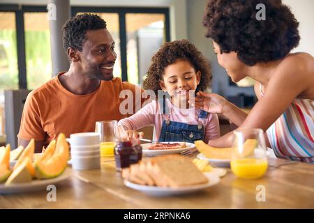 Familienschoß Mit Eltern Und Tochter Zu Hause Beim Frühstück, Das Marmelade Auf Brot Am Tisch Verteilt Stockfoto