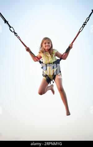 Bungee-Jumping auf Trampolin. Kleines Mädchen hüpft auf Bungee-Jumping im Vergnügungspark auf Sommerferien. Kind fliegt über den blauen Himmel, während es weiterspringt Stockfoto