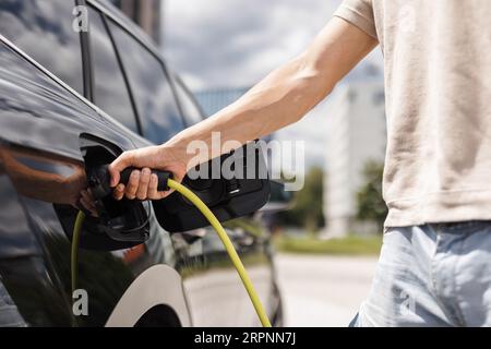 Mann, der eine Steckdose öffnet und ein Ladegerät in ein schwarzes Elektroauto an eine EV-Ladestation in der Stadtstraße stellt Stockfoto