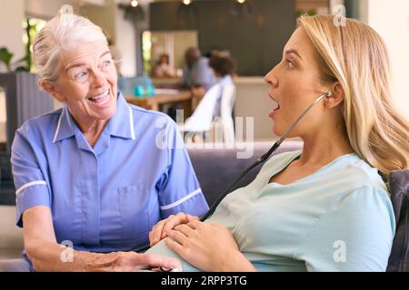 Ältere Hebamme Mit Stethoskop Besucht Schwangere Frau Zu Hause Mit Familie Im Hintergrund Stockfoto