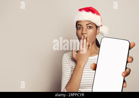 Weihnachtskonzept. Nette, überraschte Frau im Weihnachtsmann, die ein Smartphone mit leerem weißen Bildschirm und Mockup-Banner hält Stockfoto