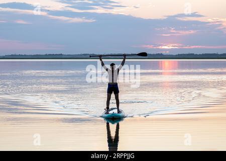 Seitenansicht Silhouette eines alleinstehenden männlichen Paddelbretts bei Sonnenuntergang. Paddeln in der Bucht. Während des Sonnenuntergangs paddelt ein junger Mann mit Stand-Up-Paddel Stockfoto