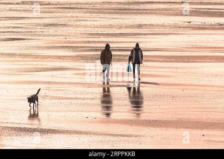 Bei Ebbe am Strand von Fistral in Newquay in Cornwall, Großbritannien, ist das Abendlicht eine Silhouette, die Menschen umspielt, die bei Ebbe am Strand von Fistral spazieren. Stockfoto