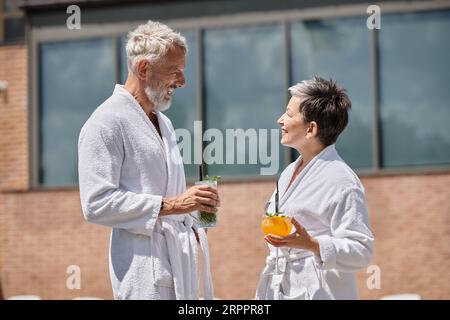 Glückliches reifes Paar in weißen Bademänteln mit Cocktails am Pool, Wellness-Retreat im Resort Stockfoto