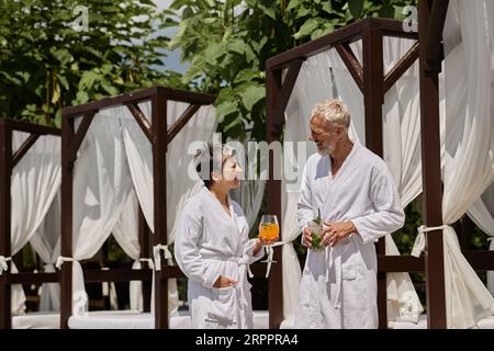 Glückliches reifes Paar in weißen Bademänteln, das Cocktails hält und sich in der Nähe des Pavillons und des Wellnessbereichs unterhalten kann Stockfoto